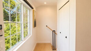 Un couloir doté d’un plancher en bois, de murs blancs, d’une fenêtre donnant sur des arbres et d’une porte avec une poignée noire.