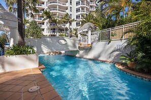 A swimming pool with a waterfall feature, surrounded by palm trees and a multi-story building with balconies. - One Bedroom Mediterranean Escape at Burleigh (Gold Coast)