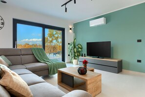 Een moderne woonkamer met een bank, een salontafel, een televisie en een groot raam met uitzicht op bomen.