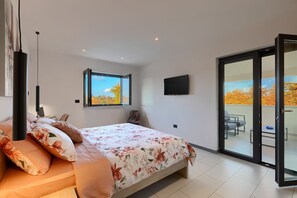 Een slaapkamer met een bed, een televisie, een raam met uitzicht op herfstbomen en een schuifdeur naar een balkon.