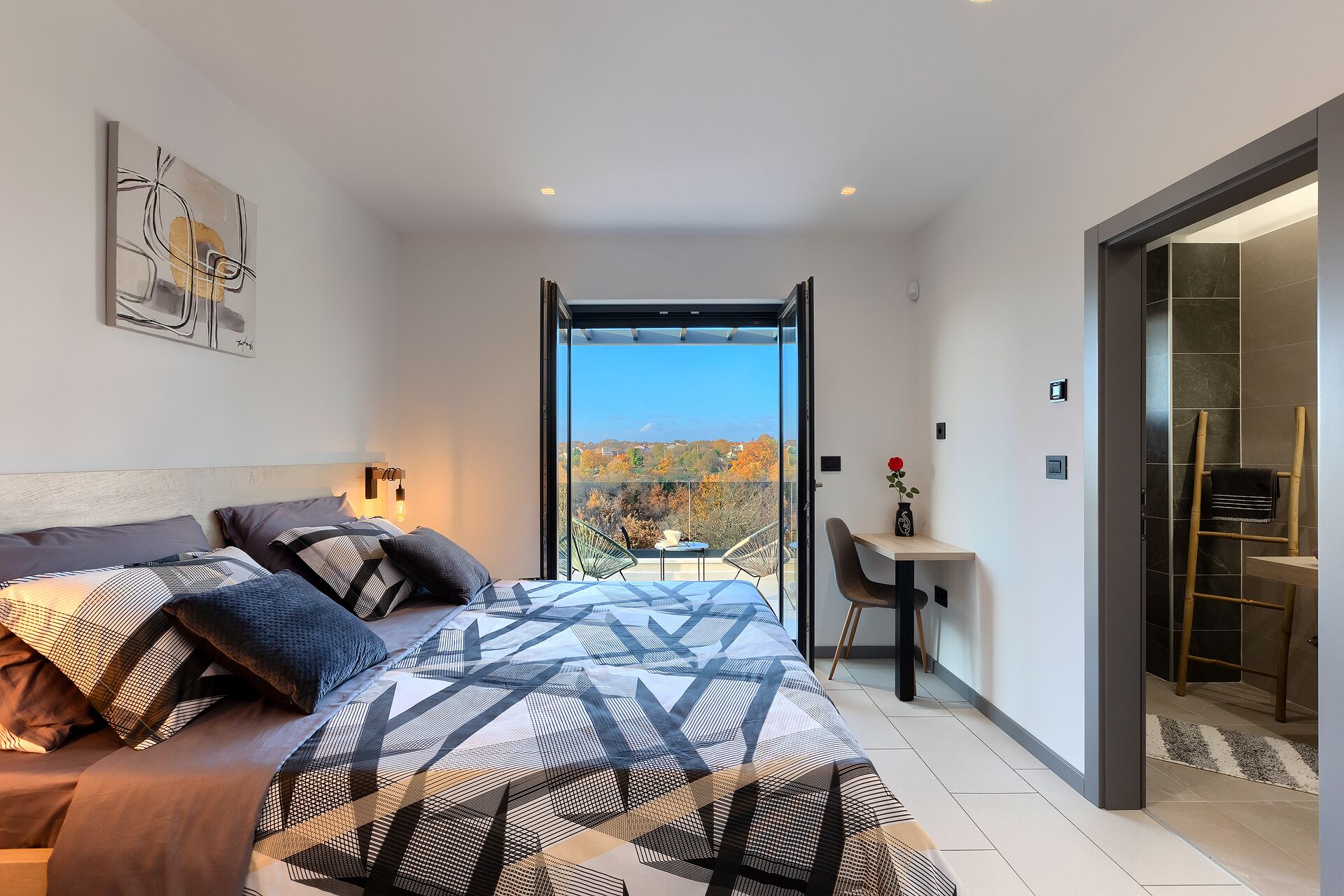 A modern bedroom with a large bed, a view of autumn foliage, a small table with a vase, and a bathroom with a towel rack.