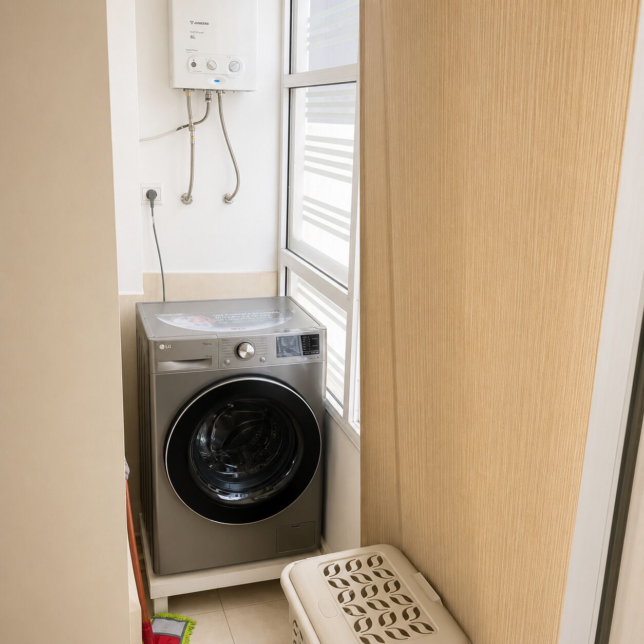 A compact laundry room with a front-loading washing machine, a wall-mounted heater, and a window with blinds.