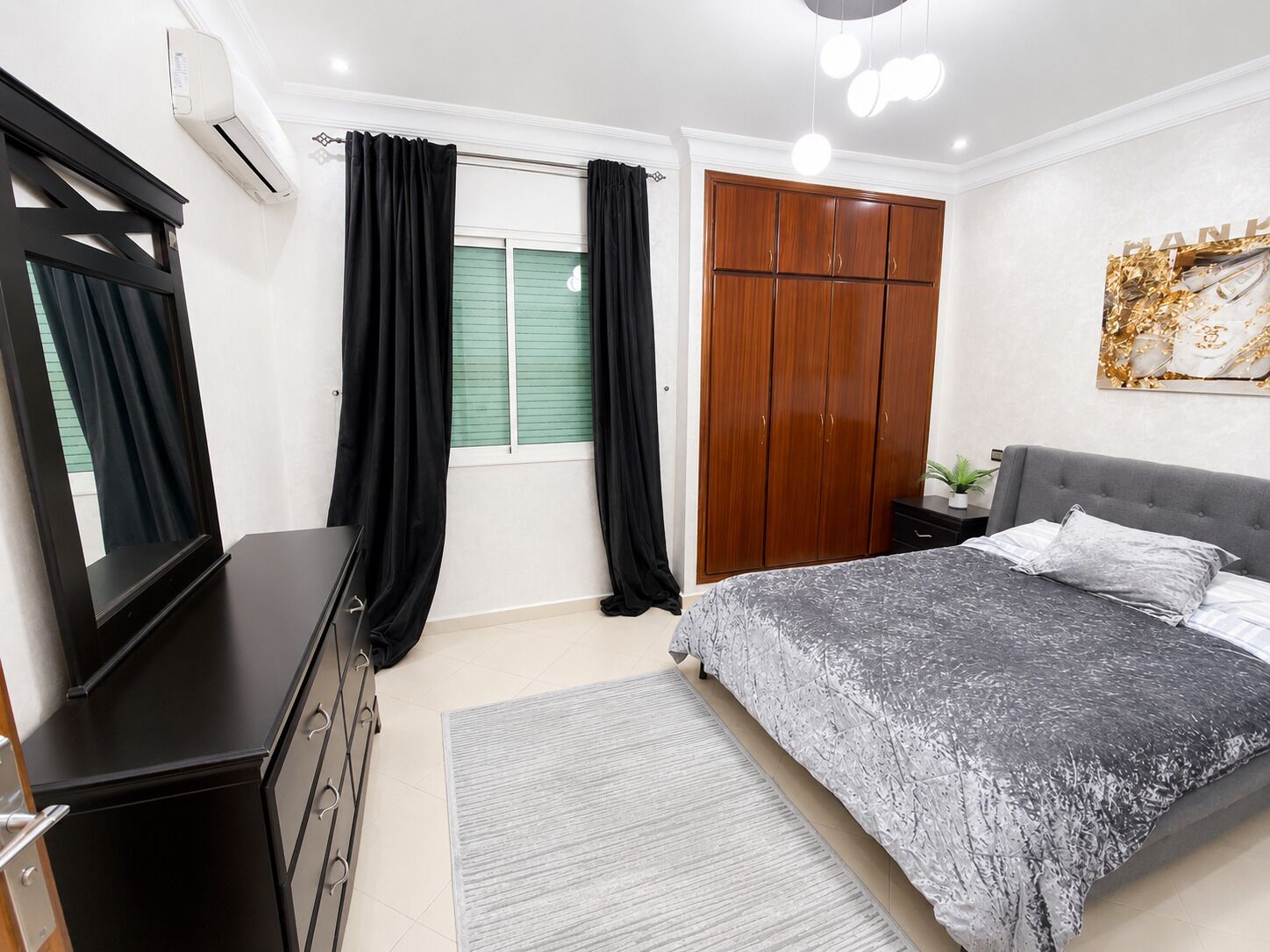 Ein Schlafzimmer mit Bett, Kommode, Kleiderschrank, einem Fenster mit Vorhängen und einer Deckenleuchte.