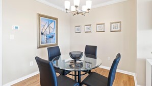 A dining area with a round glass table, four black chairs, a chandelier, and framed artwork on the wall. - Coastal Condo w/ Panoramic Water Views (St. Petersburg)
