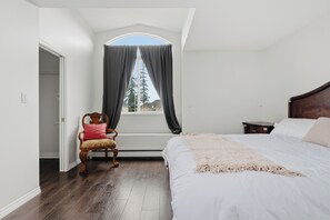 Ein Schlafzimmer mit einem großen Bett, einem Sessel, einem Nachttisch, einem Fenster mit Vorhängen und Blick auf einen Baum.