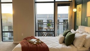 A bedroom with a large window, a bed with white bedding, a bedside table with a book and cup, and a balcony with a table and chairs. - Shaw 2BR Penthouse | Monument Views, Balconies & Rooftop (washington)