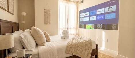 A bedroom with a bed, a bedside table with a lamp, a wall-mounted TV displaying various apps, and a macrame wall hanging.
