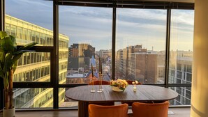 A dining area with a large window, a round table, orange chairs, and a potted plant. - Shaw 2BR Penthouse | Monument Views, Balconies & Rooftop (washington)