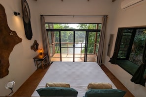 A bedroom with a bed, two pillows, a nightstand, a lamp, a large window with a view of a lake, and a wooden headboard.