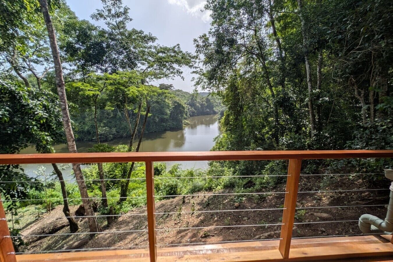 A view from a wooden balcony with a metal railing overlooking a river surrounded by dense forest.