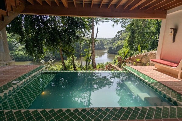 A pool with a view of a lake surrounded by trees, a wooden pergola, and a red cushioned bench.