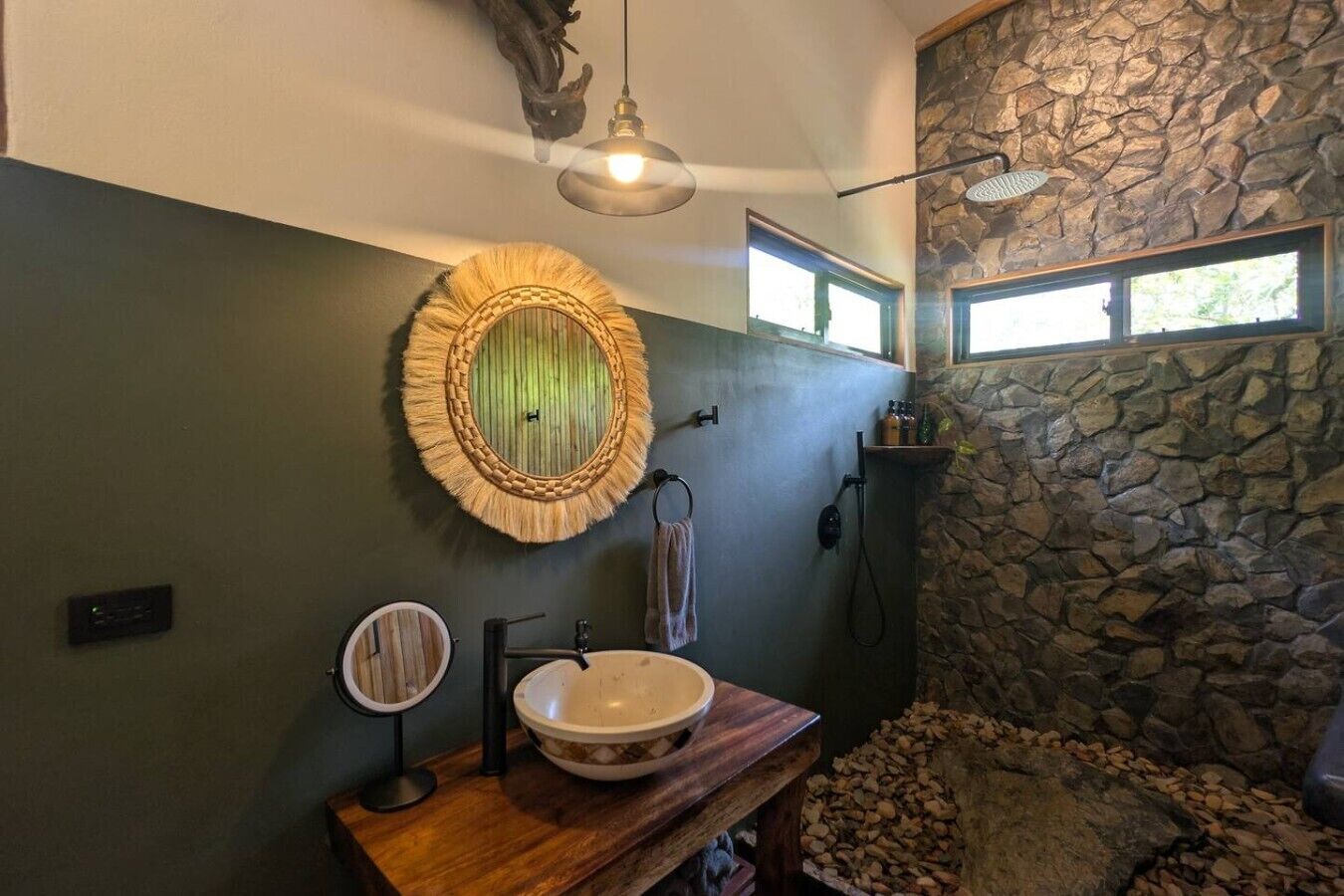 Une salle de bain avec un mur en pierre, un meuble-lavabo en bois, un miroir rond et un grand miroir rond placé au-dessus du lavabo.