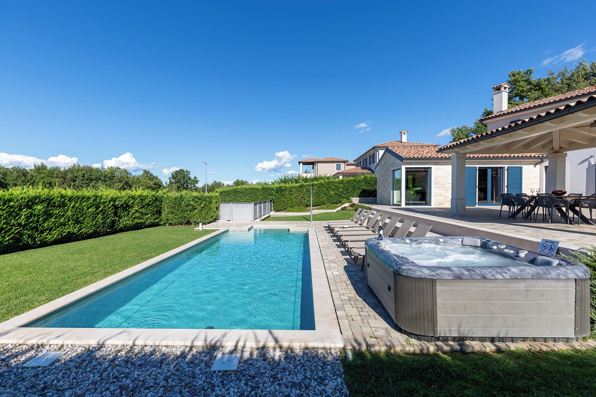 Une piscine avec un jacuzzi, un patio couvert avec des meubles d’extérieur, et une vue sur une maison au loin.