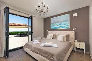 A bedroom with a large bed, a painting of waves, a chandelier, and a view of a patio with a tiled roof. - Villa Edoardo by Istrabook (Vižinada)