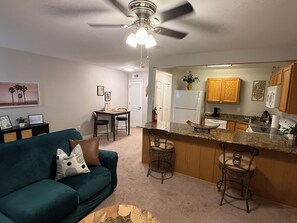 A living room with a green sofa, a ceiling fan, and a kitchen area with wooden cabinets and a granite countertop. - Cozy 1-bedroom condo in peaceful Biloxi with AC and gym access for 2 adults only (Biloxi)