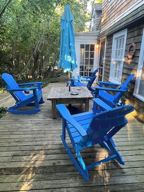 A wooden deck with a table and blue chairs, surrounded by greenery. - Historic 1932 Cherry Grove Beach House - Gardens, Sun and steps to the Beach. (Cherry Grove)