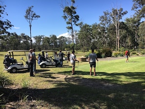 A golf course with a golf cart, people standing, and trees in the background. - River to the Sea - Jervis Bay (Myola)