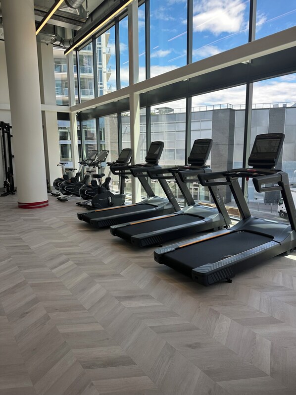 Een sportschool met loopbanden langs een muur met grote ramen, een visgraatvloer en fitnessapparatuur.