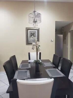 A dining area with a rectangular table, six chairs, a chandelier, a framed picture, and a potted plant. - Lakefront Oasis: Spacious 3BR/2BA West Palm Beach House (West Palm Beach)