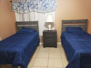 A bedroom with two single beds, a nightstand with a lamp, and a window with curtains. - Lakefront Oasis: Spacious 3BR/2BA West Palm Beach House (West Palm Beach)