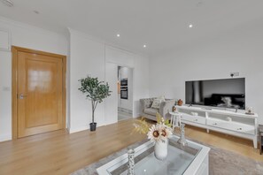 A modern living room with a wooden door, a potted plant, a glass coffee table, a sofa, a TV, and a white cabinet. - Charming 3B Apartment in Paddington Station (London)
