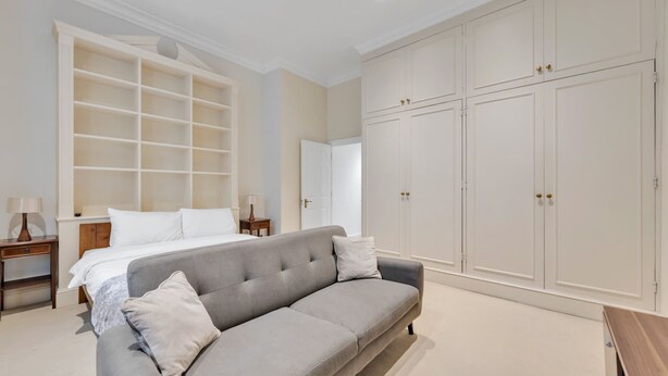 A bedroom with a grey sofa, a bed, a wardrobe, and a nightstand with lamps. - Modern Studio in Knightsbridge Greater London (London)