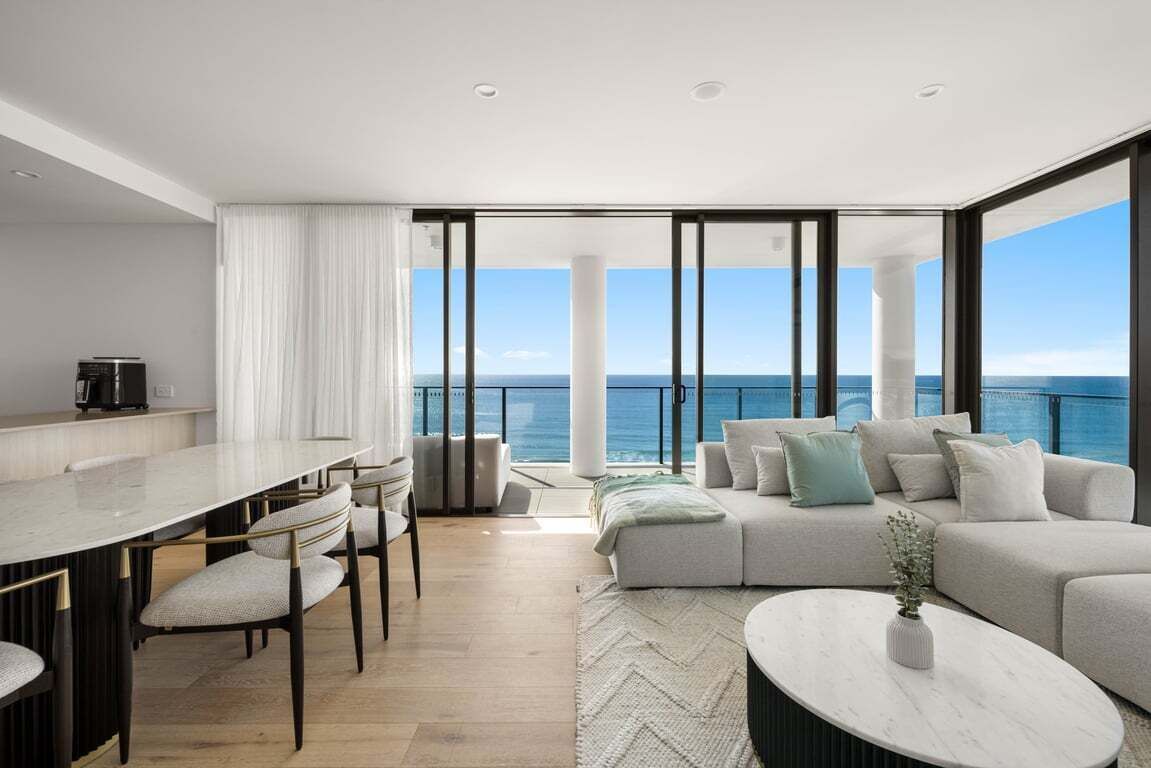 Ein modernes Wohnzimmer mit großem Fenster zum Meer, einem Essbereich mit Tisch und Stühlen sowie einer Couch mit Kissen.