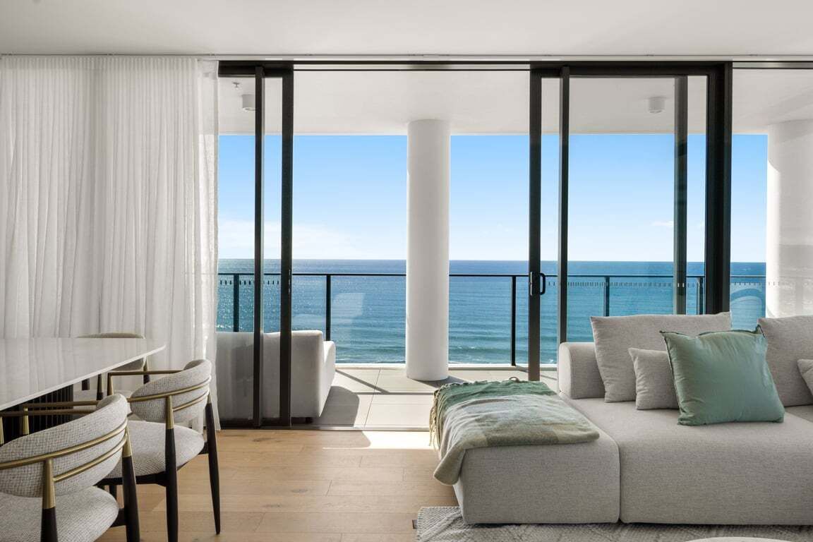 A modern living room with a balcony overlooking the sea, a dining area with a table and chairs, and a sofa with cushions.