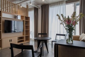 A modern living room with a flat-screen TV, wooden shelving, a dining table with chairs, a vase of flowers, and a large window with curtains. - Recoleta Prime Location | New Studio - Coworking - BBQ (Buenos Aires)