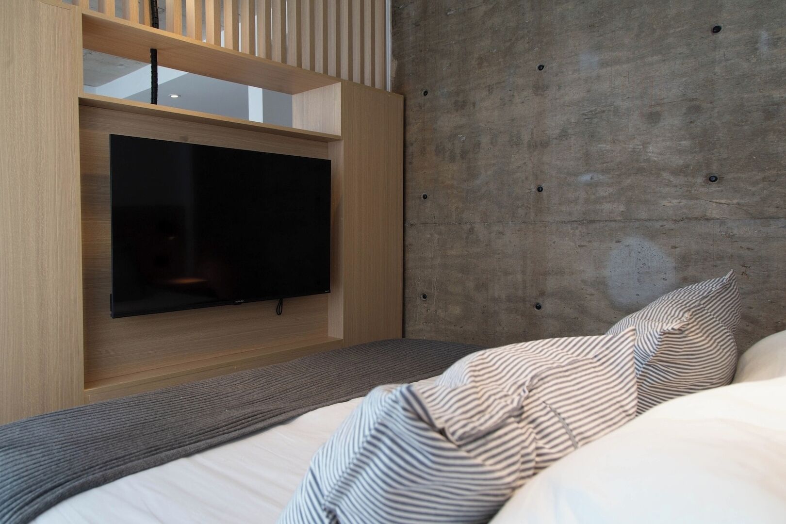 A modern bedroom with a flat-screen TV mounted on a wooden panel, a concrete accent wall, and striped bedding.