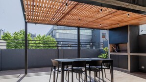 Une terrasse extérieure dotée d’une pergola en bois, d’une table avec des chaises et offrant une vue sur les bâtiments en arrière-plan.