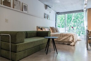 A modern room with a green sofa, a small round table, a bed with a white bedspread, and a large window with a view of trees. - Modern Recoleta Studio with BBQ Terrace and Coworking Space (Buenos Aires)