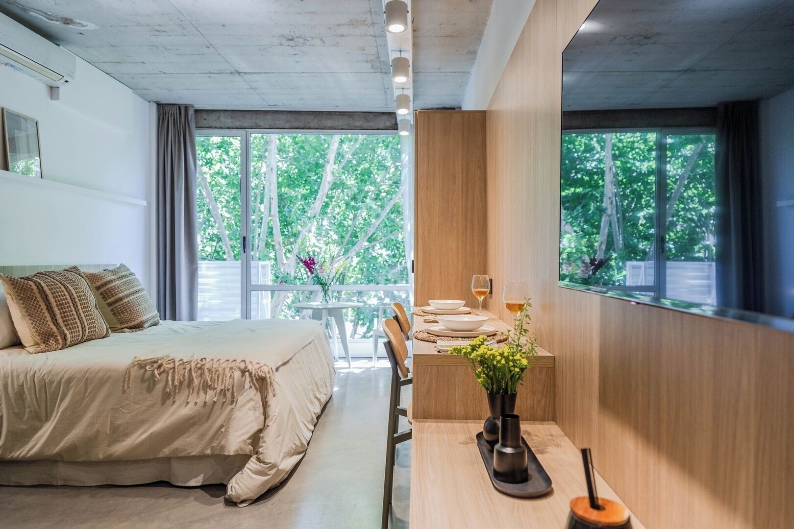 A modern bedroom with a bed, a dining table with chairs, and large windows with a view of trees.