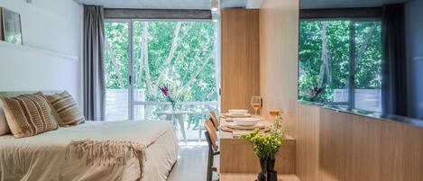 A modern bedroom with a bed, a dining table with chairs, and large windows with a view of trees.