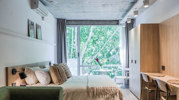 Une chambre à coucher moderne avec un lit, un canapé vert, un bureau en bois avec des chaises et un balcon donnant sur des arbres.