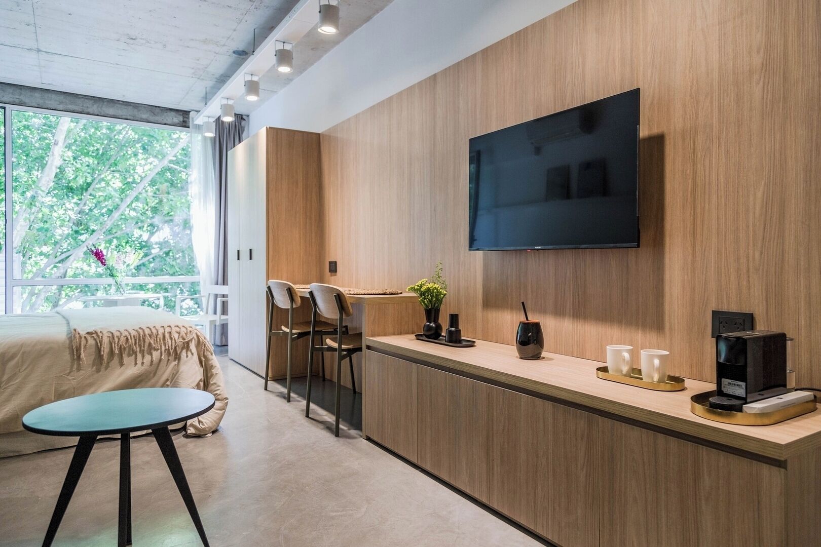 A modern hotel room with a wooden accent wall, a flat-screen TV, a small bar area with stools, and a bed with a view of greenery outside.