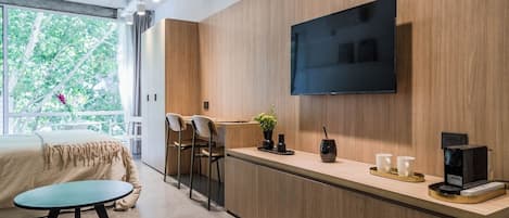 A modern hotel room with a wooden accent wall, a flat-screen TV, a small bar area with stools, and a bed with a view of greenery outside.