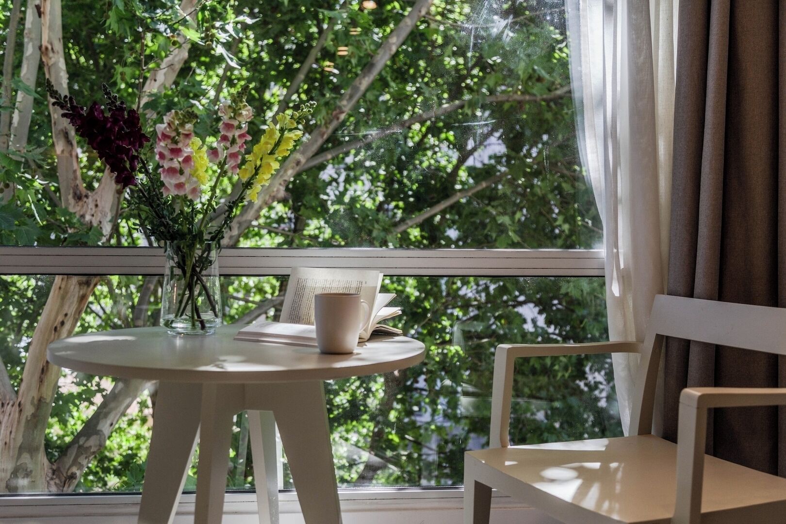 A round table with a book and a mug, a chair, and a vase with flowers on a balcony with a view of trees.