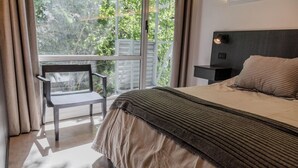 Een slaapkamer met een bed, nachtkastje, stoel en raam met uitzicht op bomen.