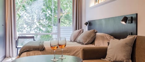 A room with a bed, a round table with two glasses of amber liquid, a headboard, and a window with curtains.