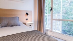 Ein Schlafzimmer mit Bett, Nachttisch, Lampe und einem Fenster mit Blick auf Grünflächen.