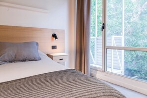 Ein Schlafzimmer mit Bett, Nachttisch, Lampe und einem Fenster mit Blick auf Grünflächen.