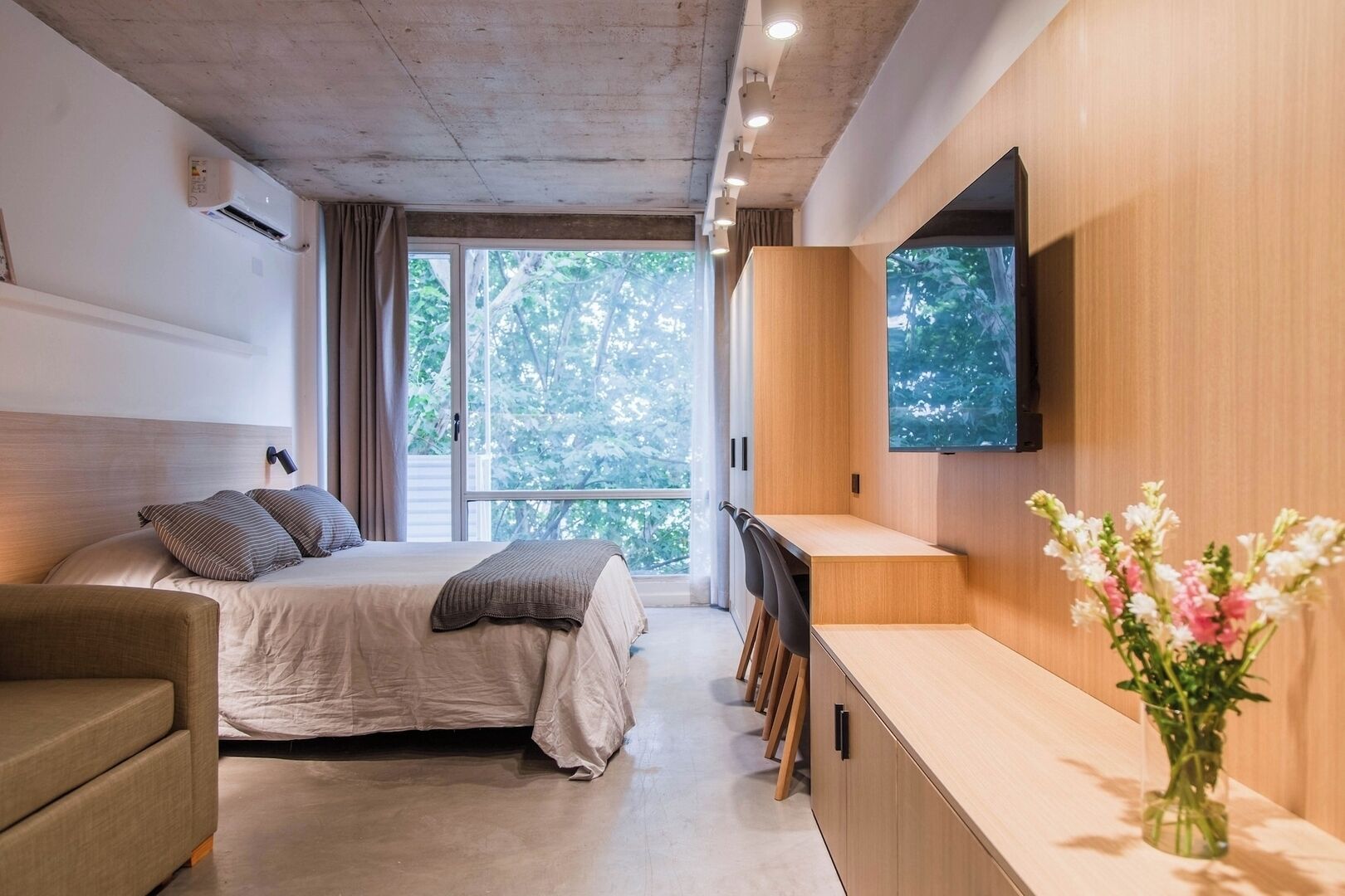 Quarto moderno com cama, mesa de cabeceira, televisão e vista para árvores.