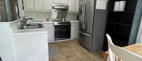 A kitchen with white cabinets, a stainless steel refrigerator, a stove, and a dining area with a wooden table and white chairs.