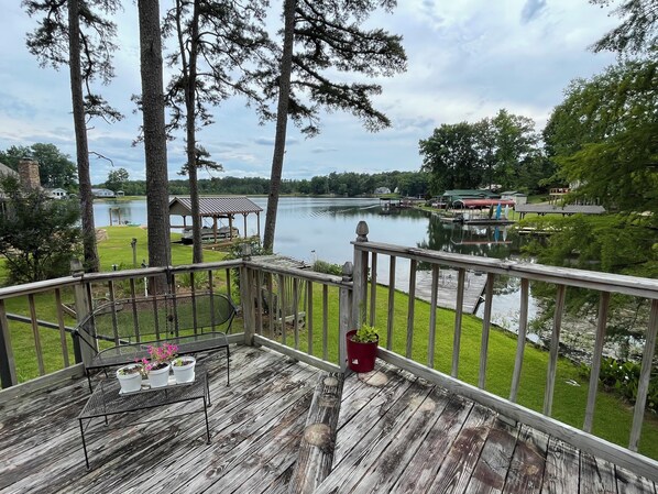Terraza de madera con barandilla que da a un lago, rodeada de árboles y un pabellón.