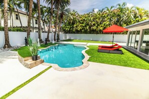 Una piscina con una sombrilla roja, rodeada de césped artificial y palmeras.