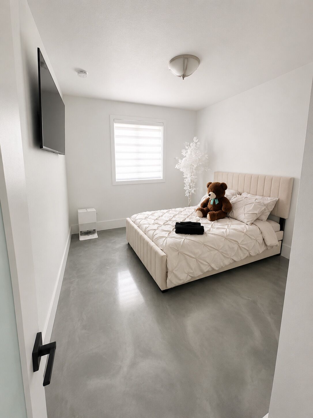 A bedroom with a bed, a teddy bear, a TV, a window, and a white wall.