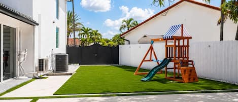 A backyard with a swing set and slide, a white fence, and a building with a balcony.