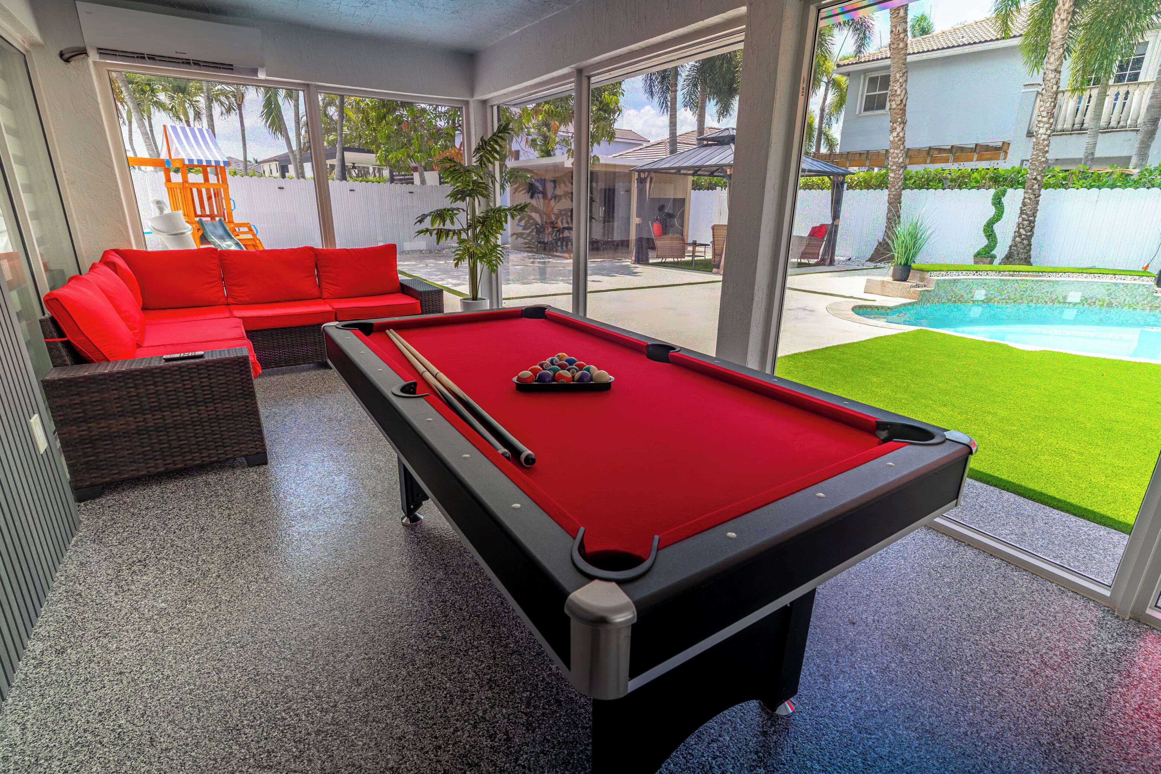 A pool table with a red felt surface, a red sofa, and a glass-enclosed area with a view of a backyard.