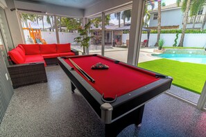 A pool table with a red felt surface, a red sofa, and a glass-enclosed area with a view of a backyard.